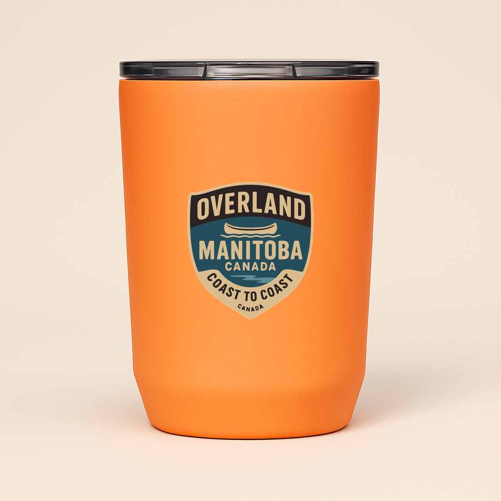 A woman in an orange shirt and green shorts stands next to a blue canoe labeled 'Overland Manitoba Coast to Coast' in a scenic outdoor setting.